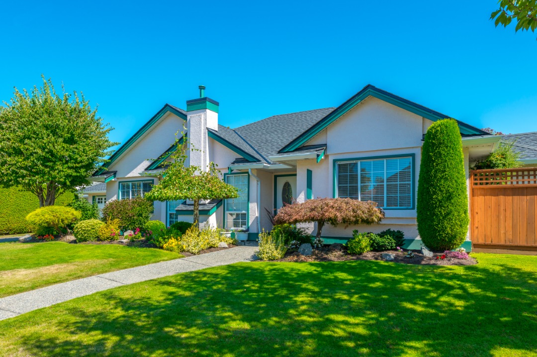 Light-colored suburban house with green trim, landscaped garden beds, shaped bushes, and healthy green front lawn.