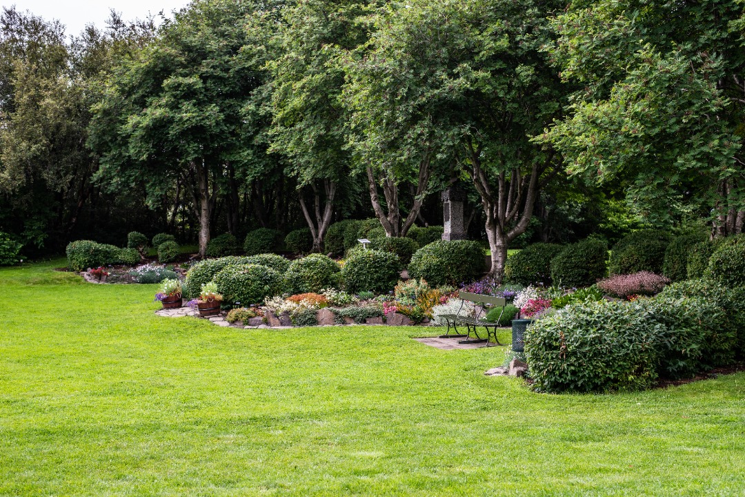 Park-style garden with trimmed hedges, flower beds, benches, and dense tree coverage creating a shaded, peaceful setting.