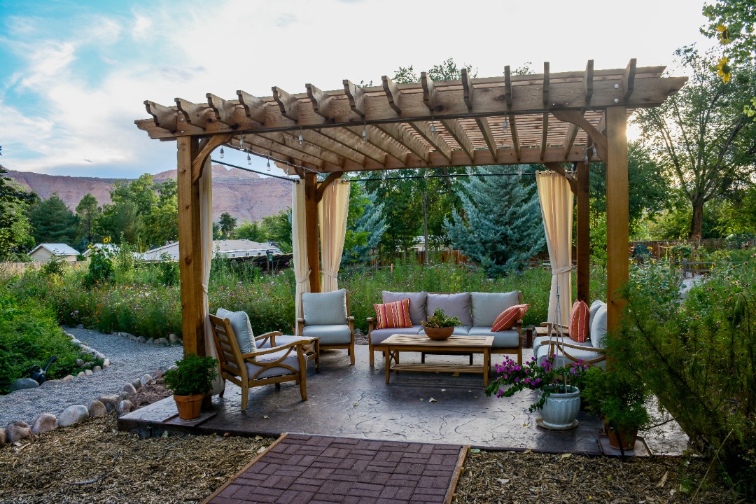 Wood pergola shades cozy outdoor seating with pillows and potted plants, surrounded by lush landscaping and trees.