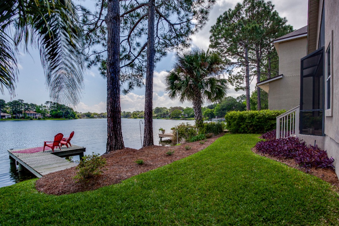  Lakeside backyard with palm trees, manicured grass, and red chairs on a wooden dock overlooking the calm water.