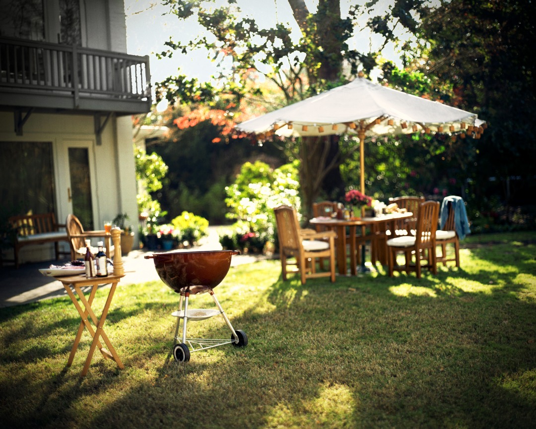 Backyard patio with barbecue grill, wooden dining set, and umbrella surrounded by lush plants and soft afternoon sunlight.