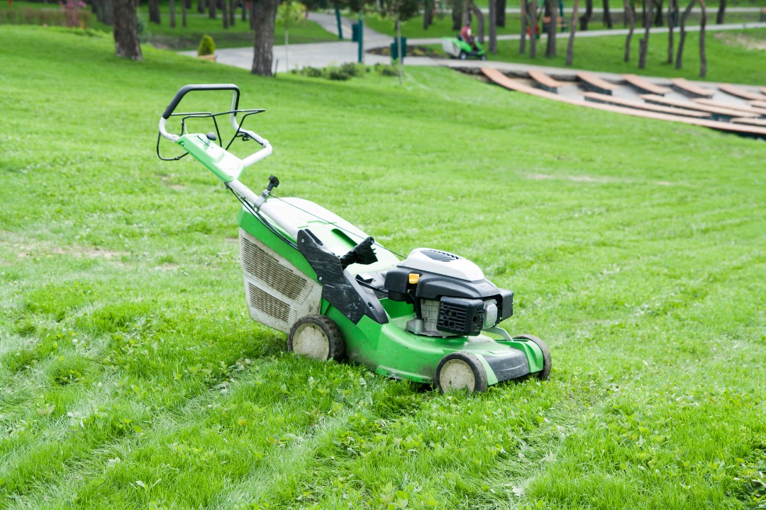 Green push lawn mower sits on freshly cut grass in a park-like setting with trees and walkways in the background.
