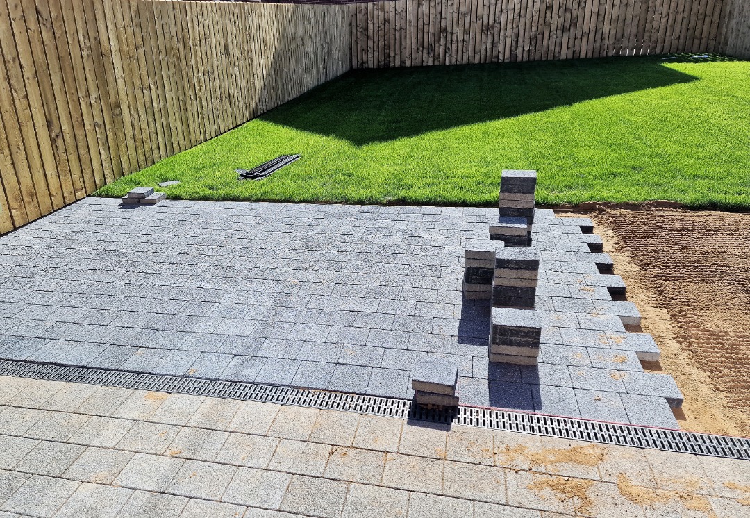 Backyard patio installation in progress with stacked pavers, drainage channel, and fresh sod against a wooden privacy fence.