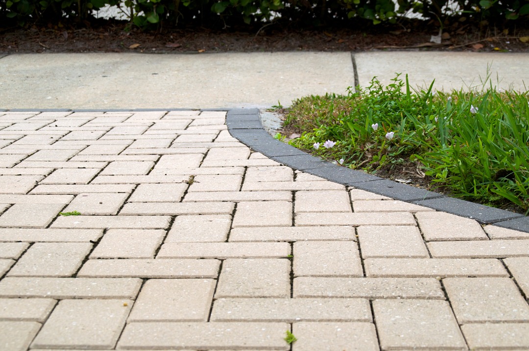 Curved brick paver walkway bordered by grass and edged with contrasting stone along a sidewalk and landscaped area.