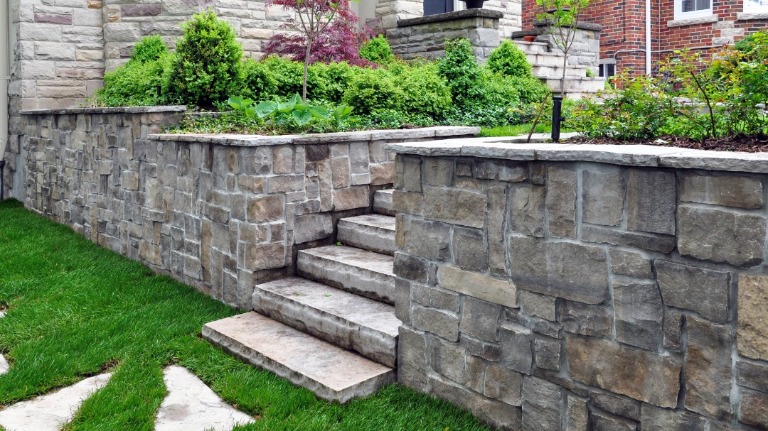 Tiered stone retaining wall with wide steps, lush green shrubs, and formal landscaping in front of a brick home.